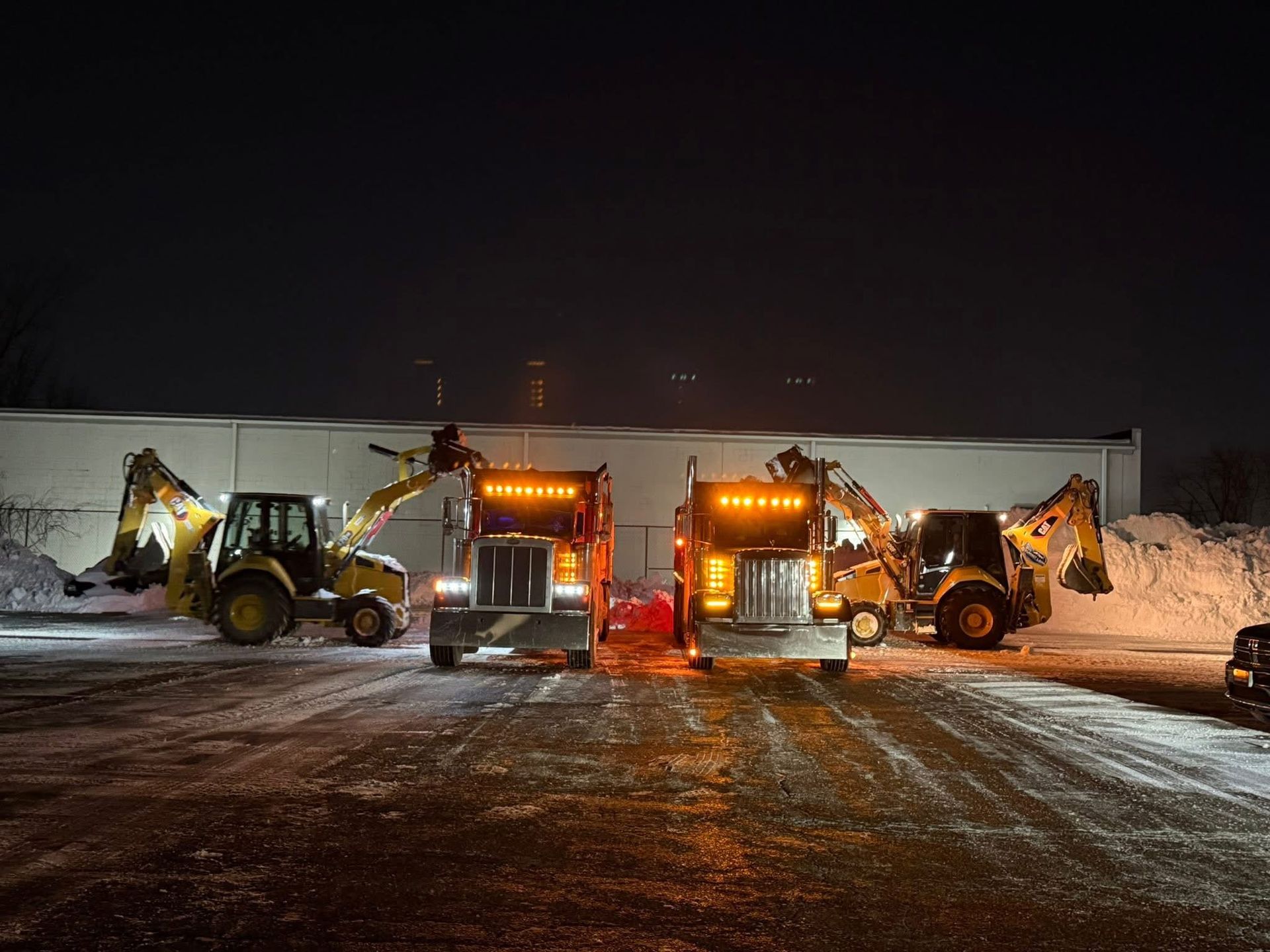 Two snowplow trucks and excavators lined up in a snowy lot at night.