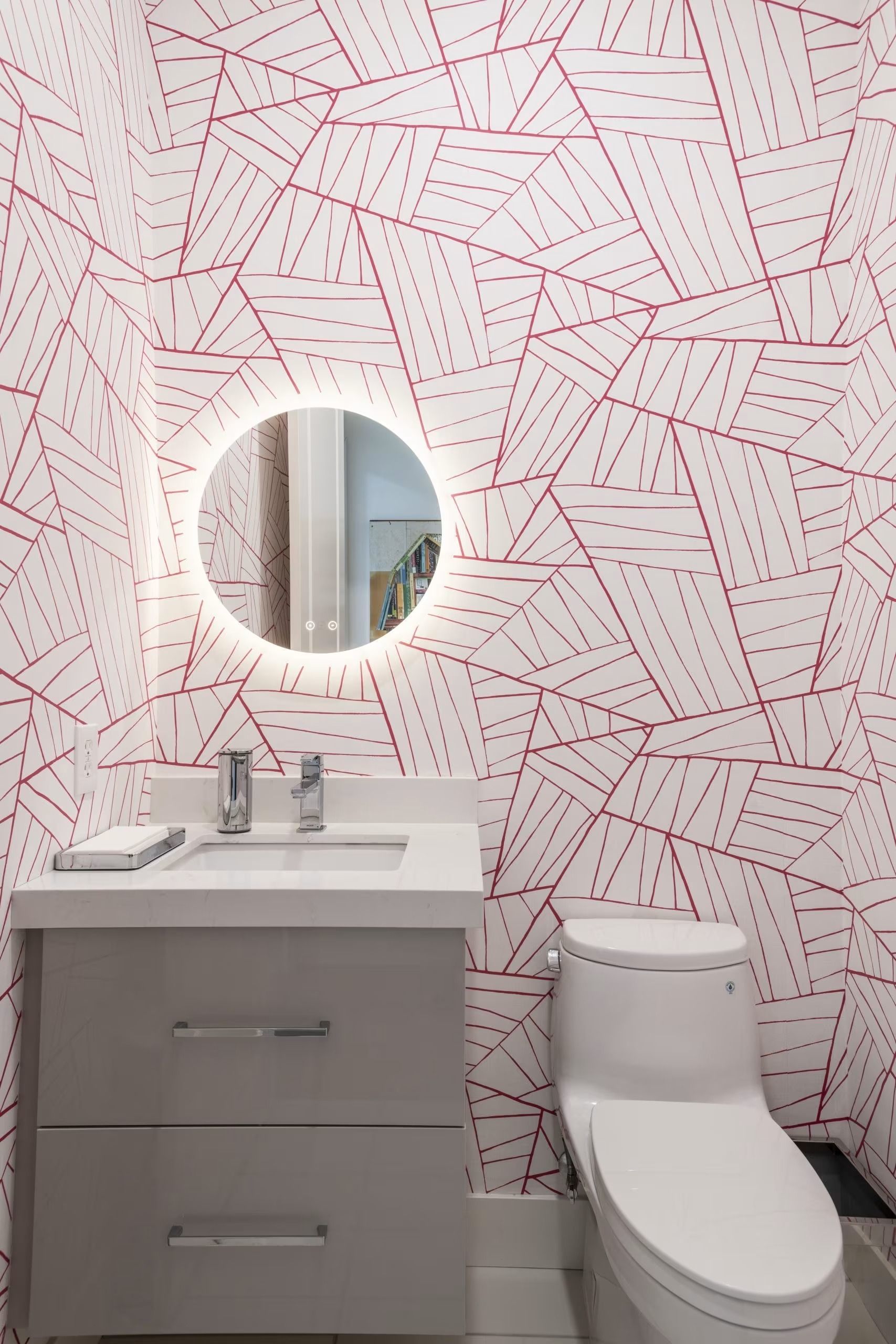 Modern bathroom with gray vanity, round lighted mirror, white toilet, and pink patterned wall.