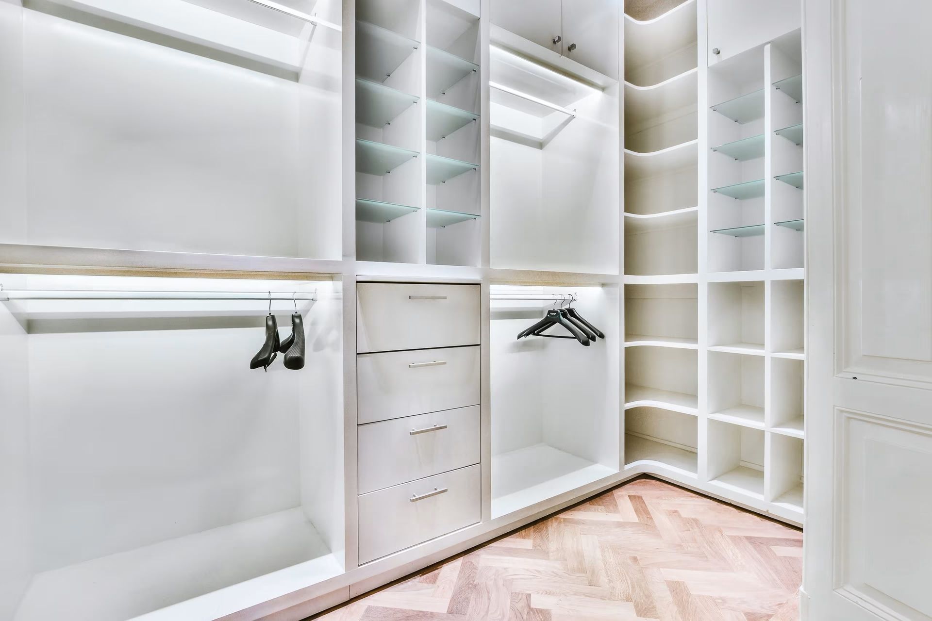Bright white walk-in closet with built-in shelves, drawers, hanging rods, and herringbone wood floor.