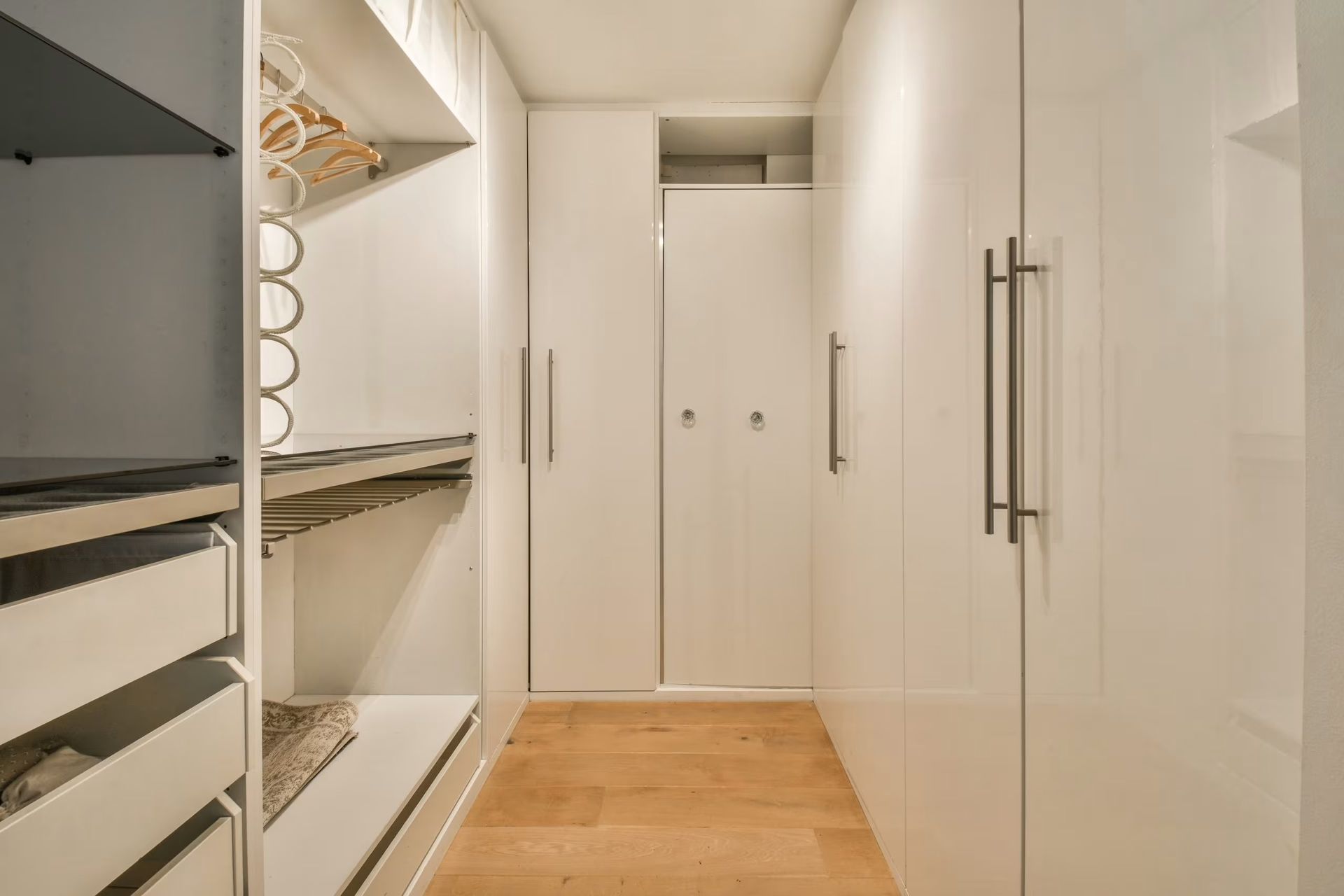 Narrow walk-in closet with white shelving, hanging rods, and light wood floor.