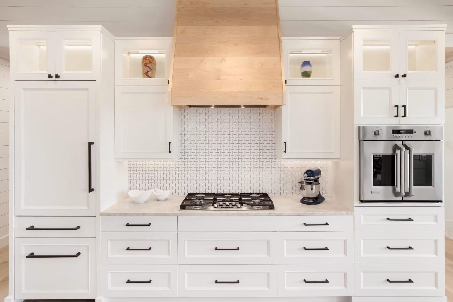 Bright white kitchen with wood range hood, mosaic backsplash, gas cooktop, and built-in stainless oven.
