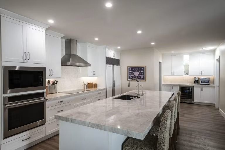 Modern white kitchen with large marble island, built-in ovens, stainless range hood, and recessed lighting