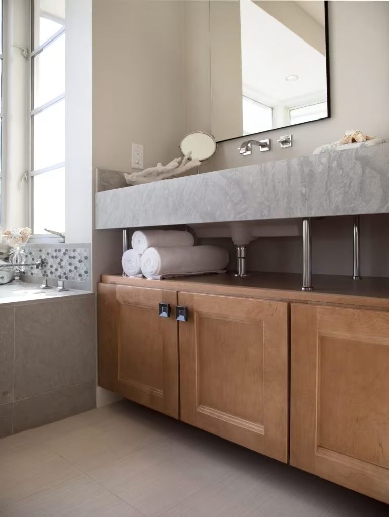 Modern bathroom vanity with wood cabinets, gray countertop, mirror, and window seat by a large window