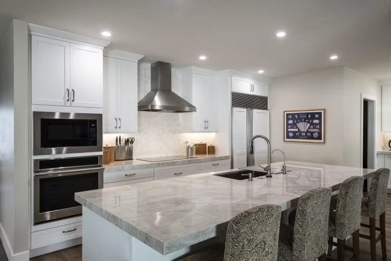 Modern white kitchen with marble island, stainless range hood, built-in oven, and four patterned barstools