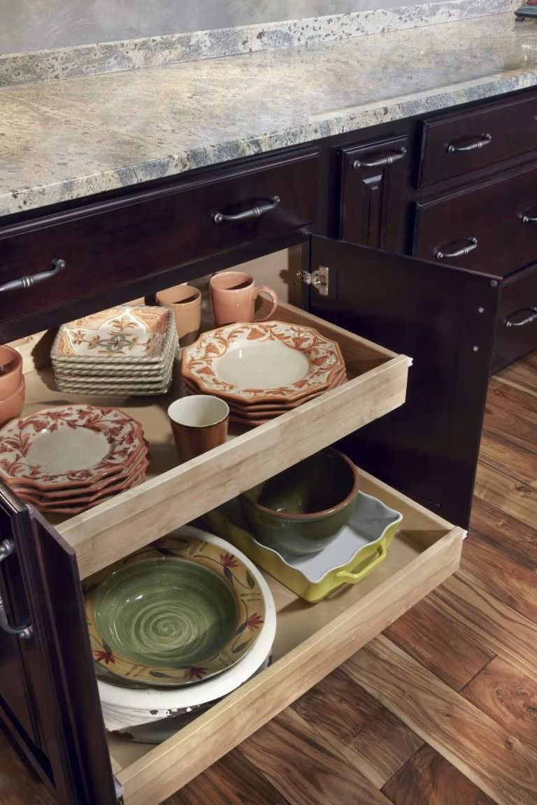 Kitchen drawer with pull-out trays holding stacked plates, bowls, and cups under a countertop.
