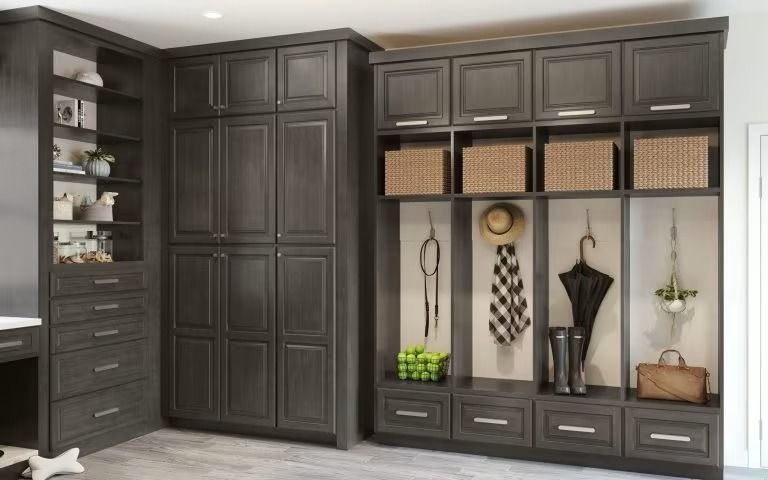 Dark built-in mudroom cabinets with open cubbies, hooks, drawers, and woven baskets in a bright room