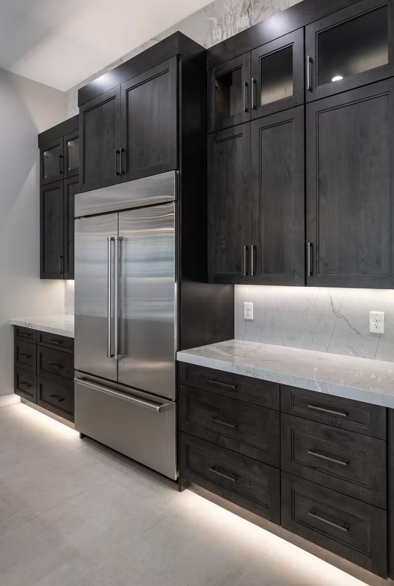 Modern kitchen with dark cabinets, stainless steel refrigerator, and white marble countertops.