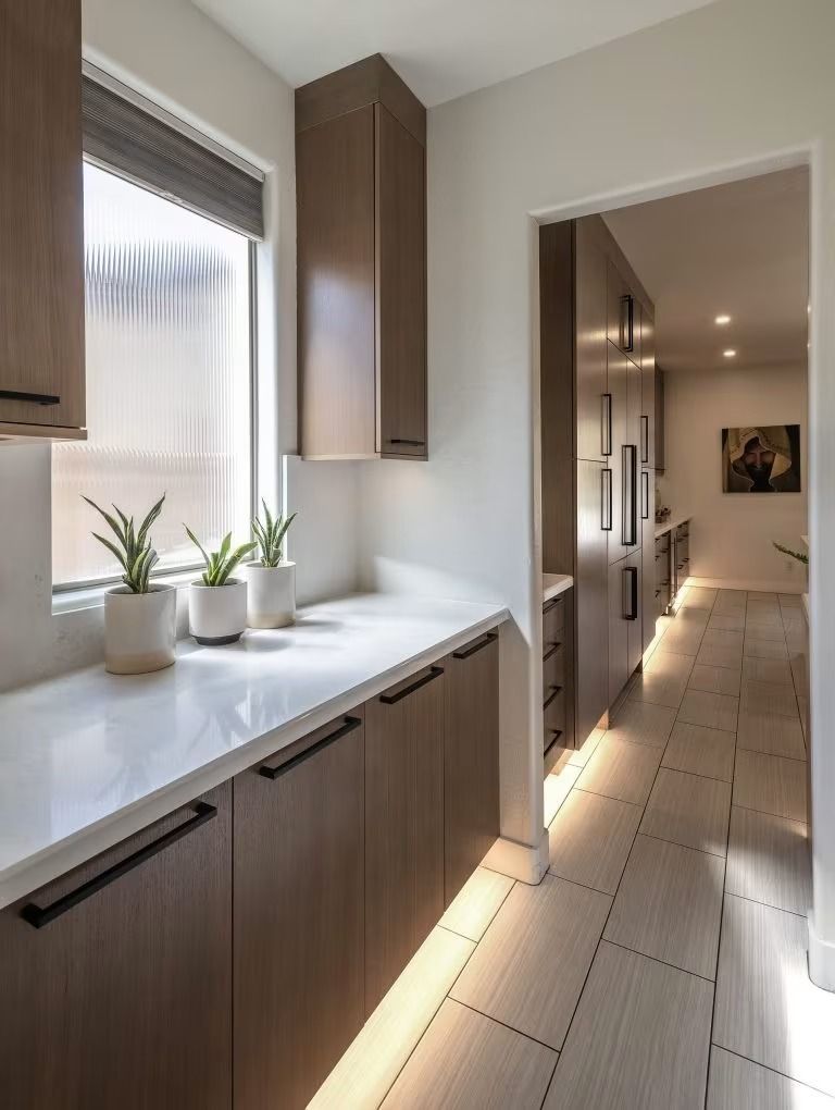 Bright modern kitchen hallway with wood cabinets, white countertops, and potted plants by a window.