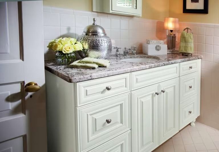 White bathroom vanity with granite countertop, flowers, towels, and decorative lamp under a window