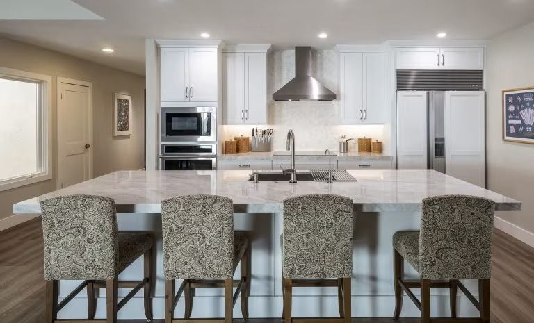Modern kitchen with island, barstools, white cabinets, stainless range hood, and built-in ovens