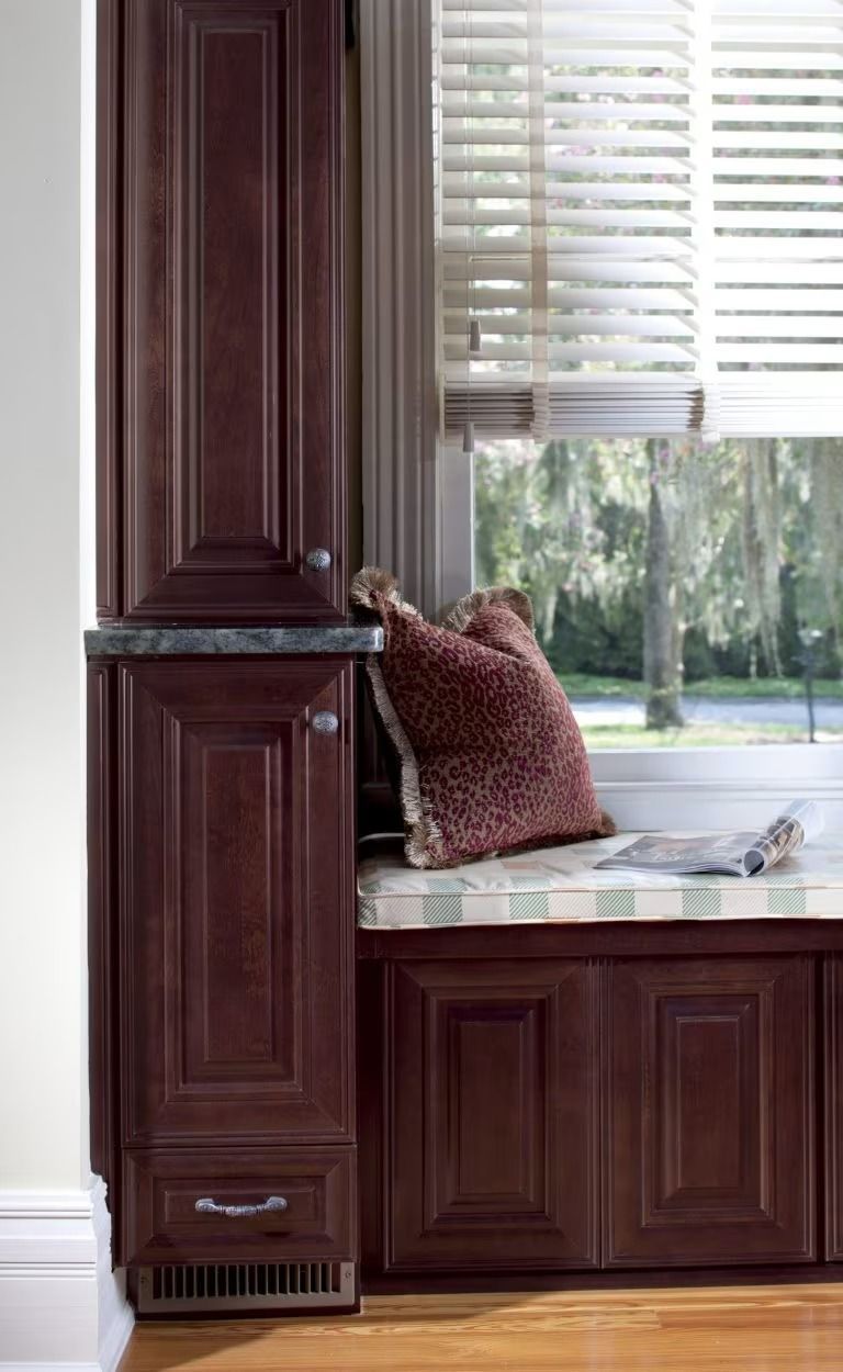 Person leaning on a kitchen counter beside dark wood cabinets and a window with blinds.