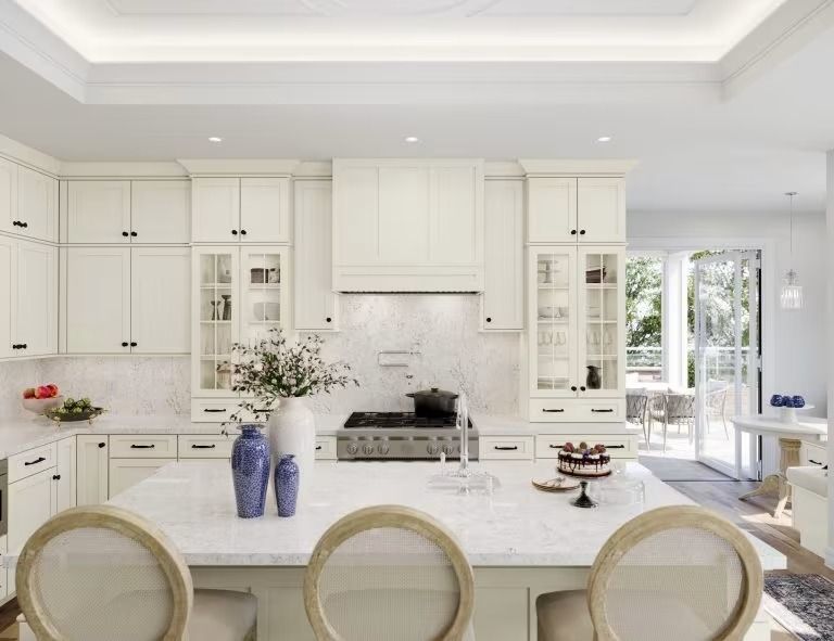 Bright white kitchen with island, rattan stools, glass cabinets, and natural light from a window and glass doors