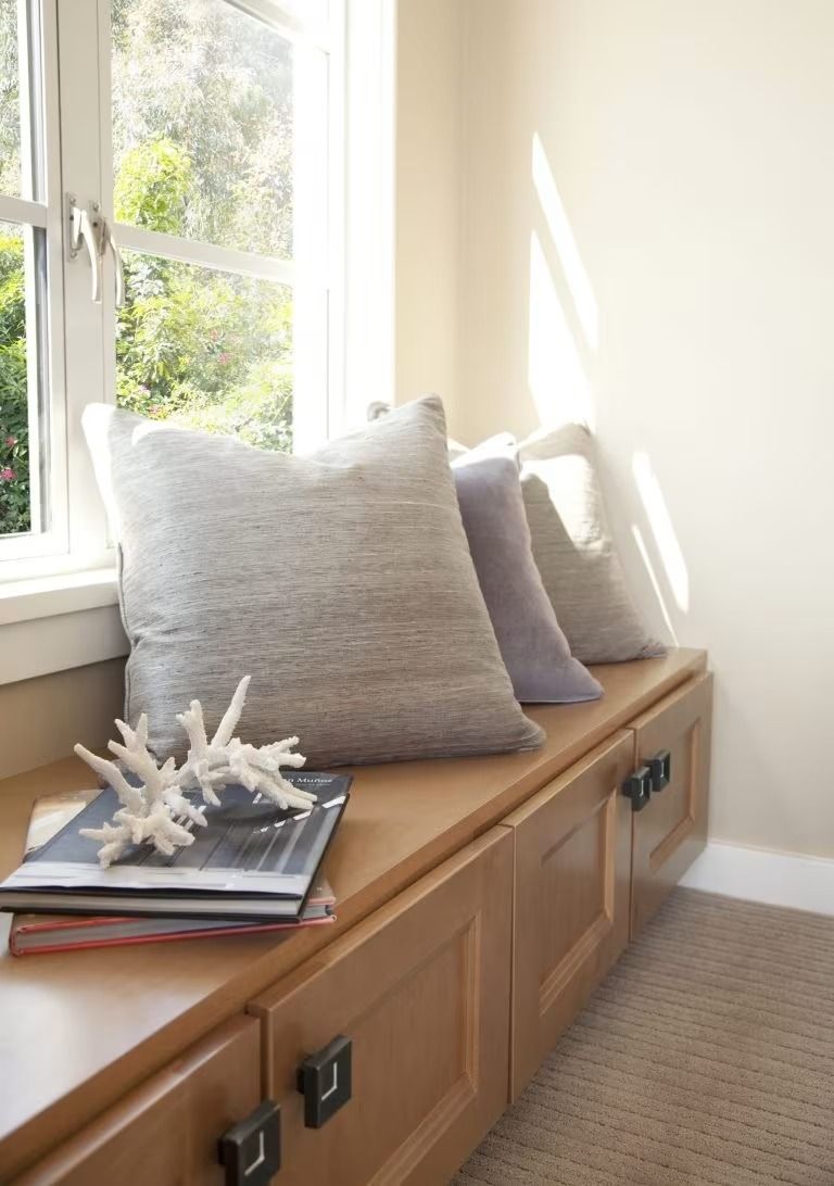 Window seat with gray pillows, a book, and a coral decoration on a wooden cabinet in a bright room