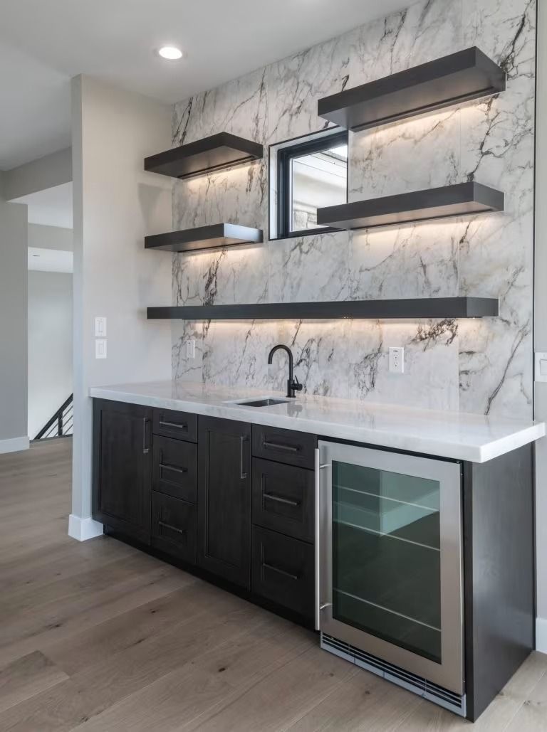 Modern kitchen wet bar with marble backsplash, dark cabinets, floating shelves, and glass-door wine fridge