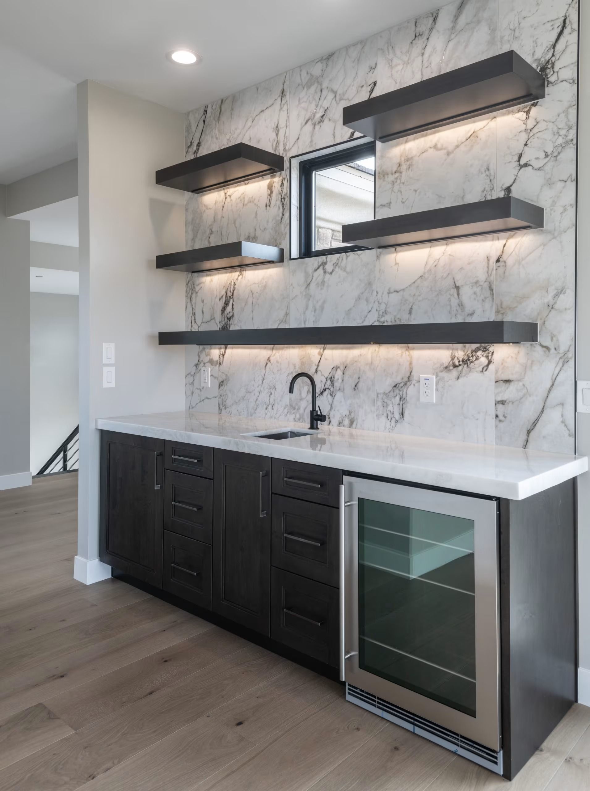 Modern kitchen wet bar with marble backsplash, floating shelves, dark cabinets, and a wine fridge