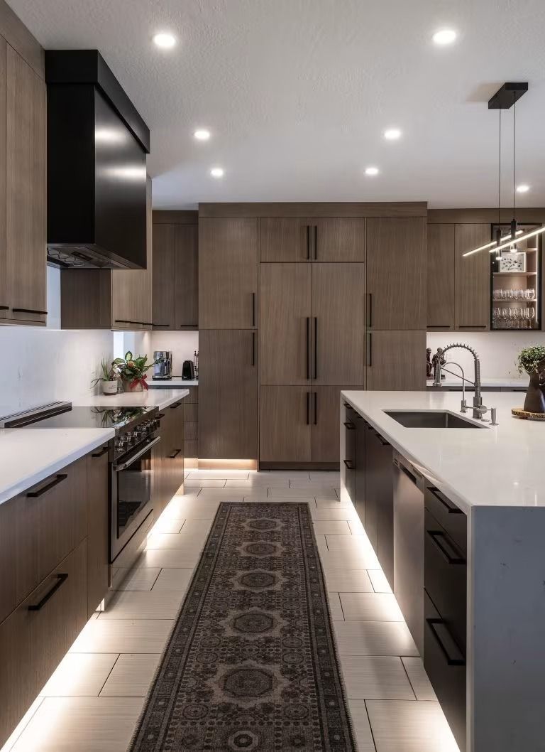 Modern galley kitchen with white countertops, dark cabinets, warm under-cabinet lighting, and patterned runner rug