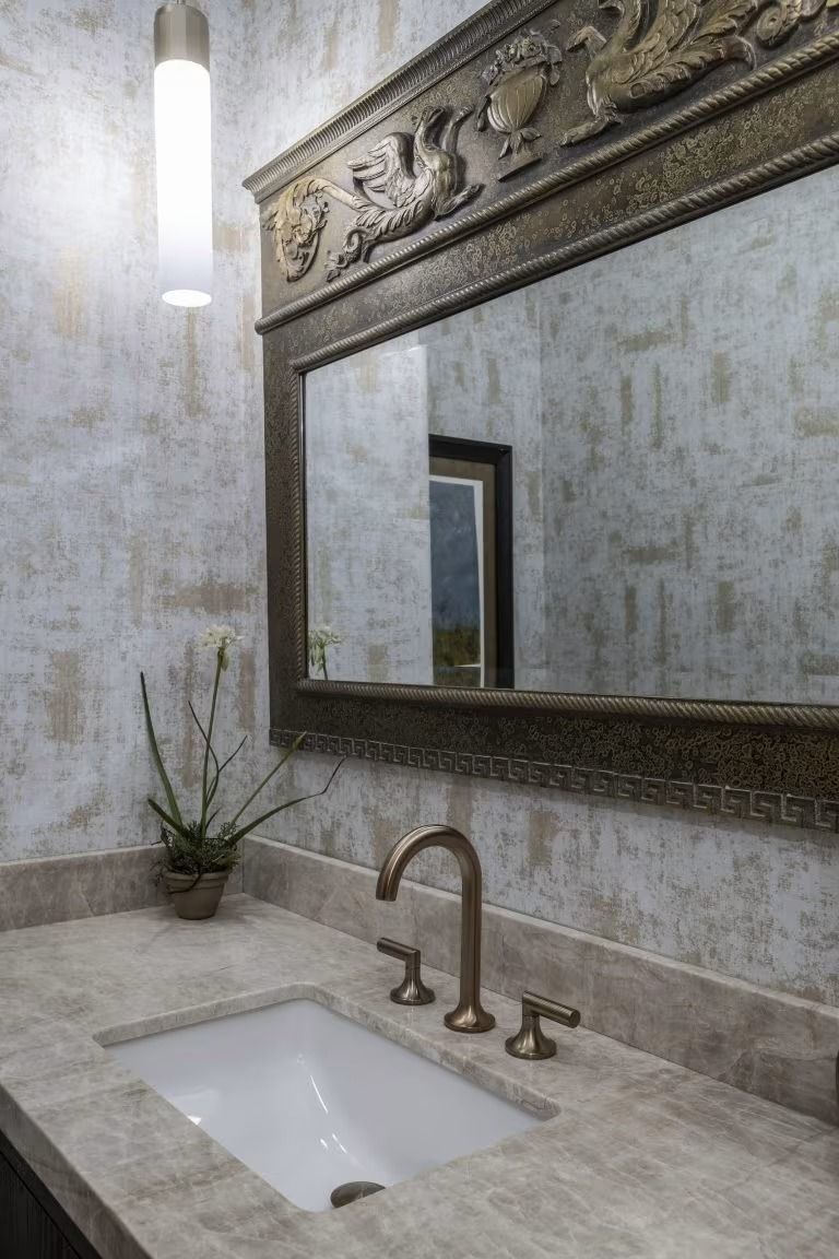 Elegant bathroom vanity with ornate mirror, brass faucet, white sink, and a lit wall sconce.