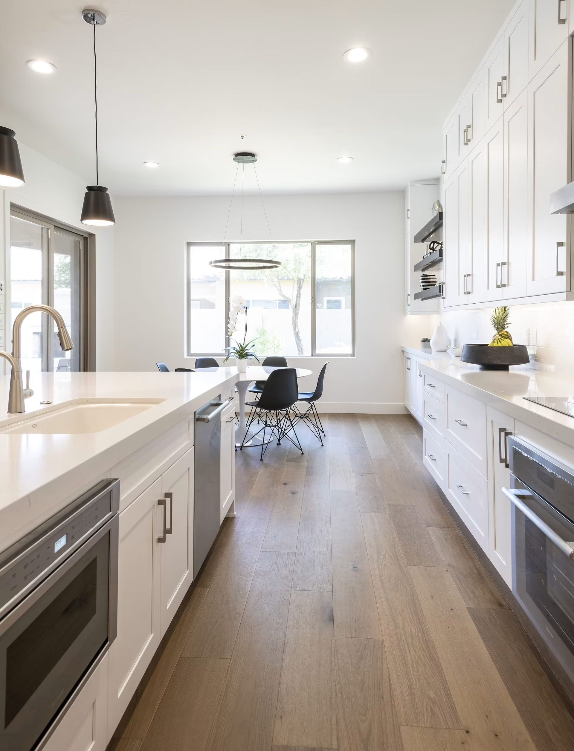 Modern white kitchen with island, stainless appliances, hardwood floor, and dining area by a bright window