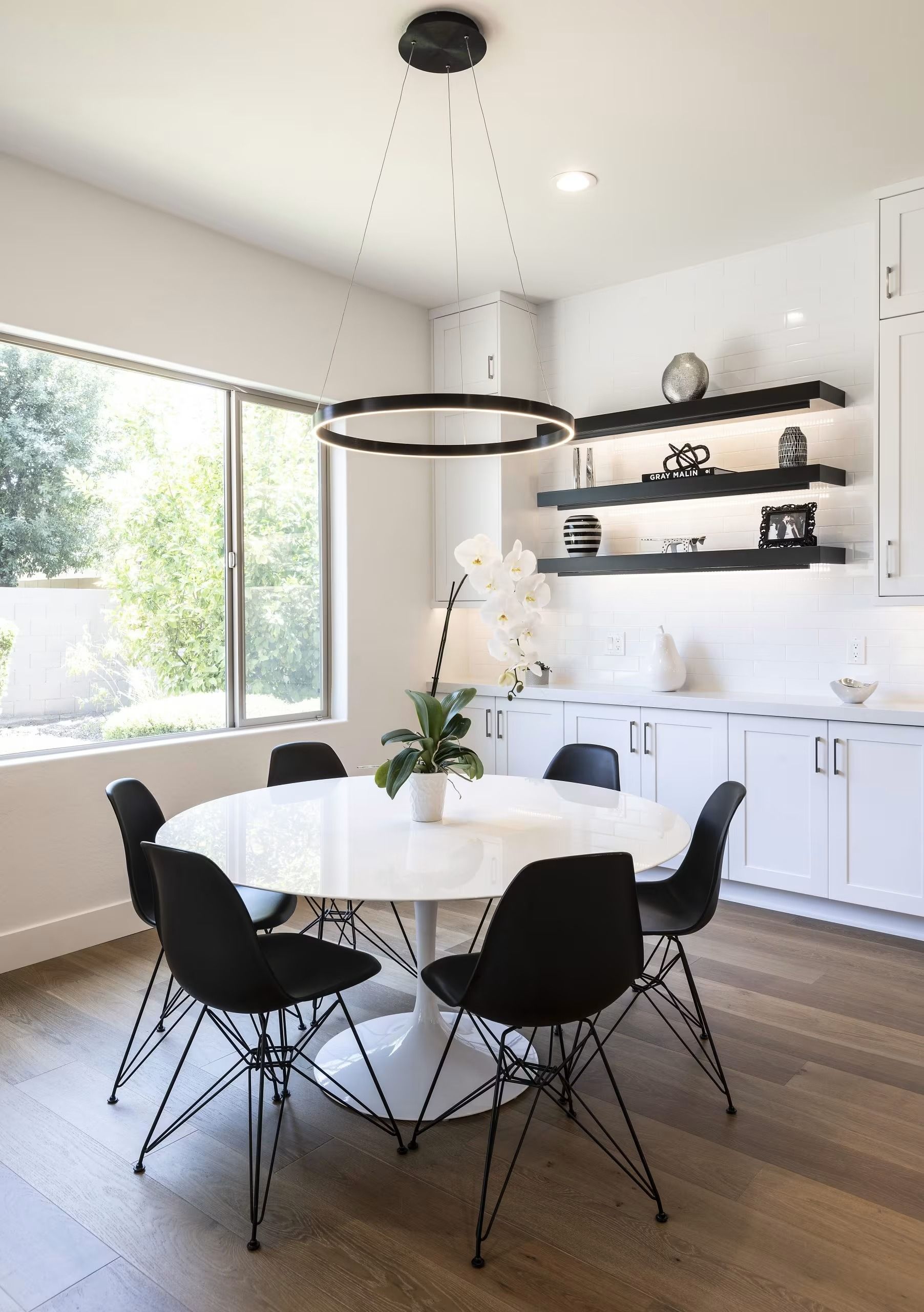 Bright modern dining room with round white table, black chairs, and wall shelves by a large window