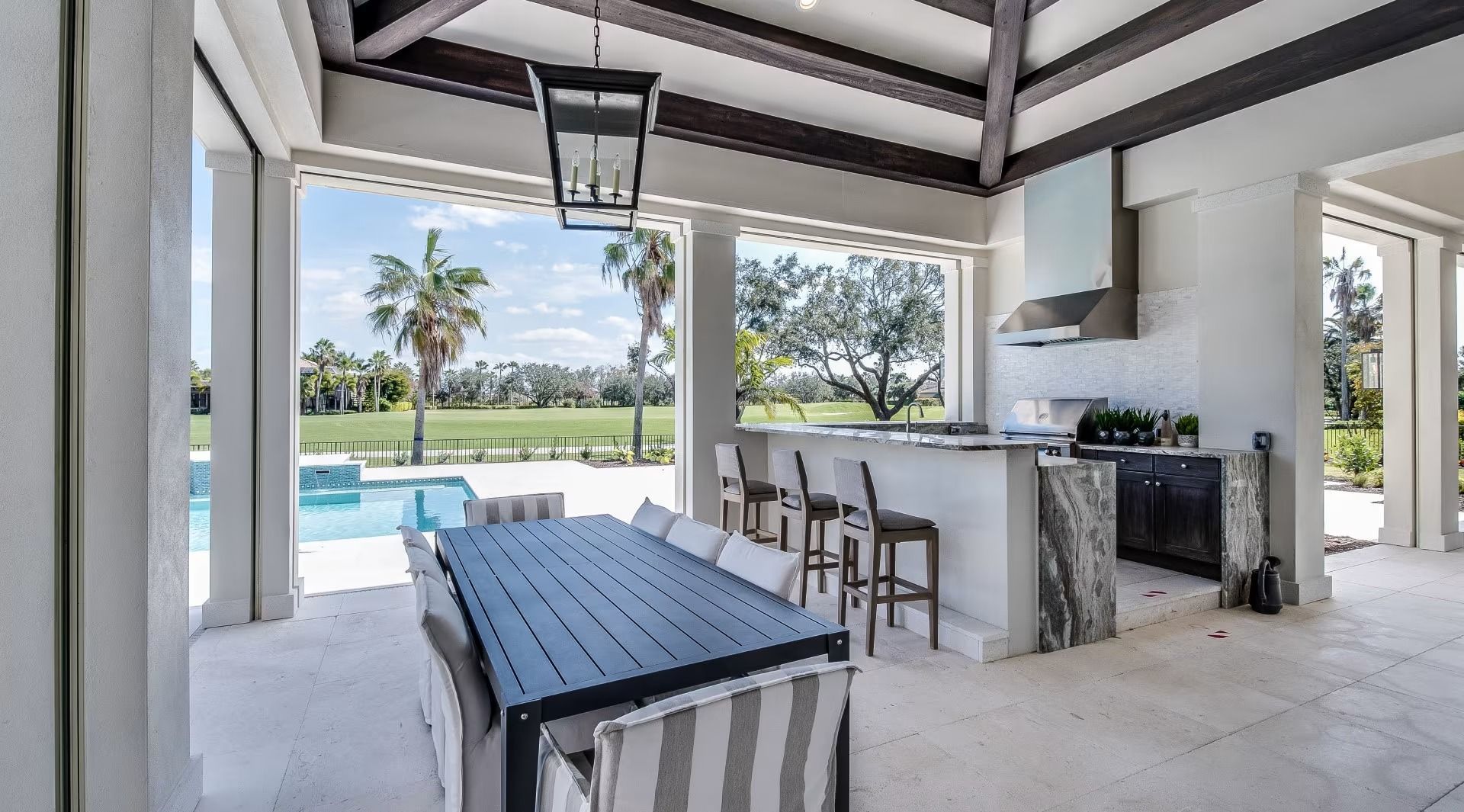 Covered outdoor patio with dining table, bar stools, and grill overlooking a pool and grassy yard