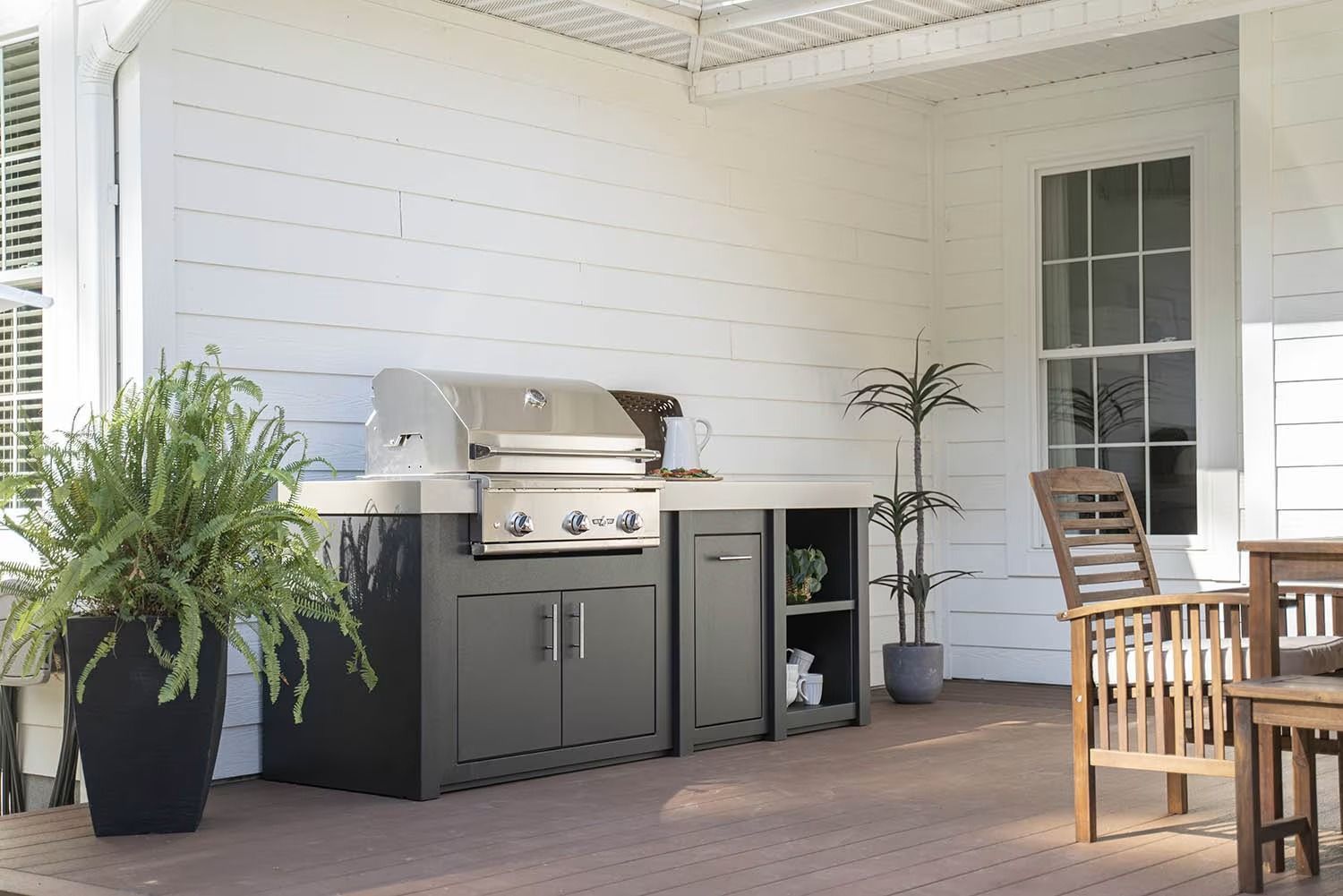 Covered patio with stainless grill, potted plants, and wooden outdoor seating beside a white house.