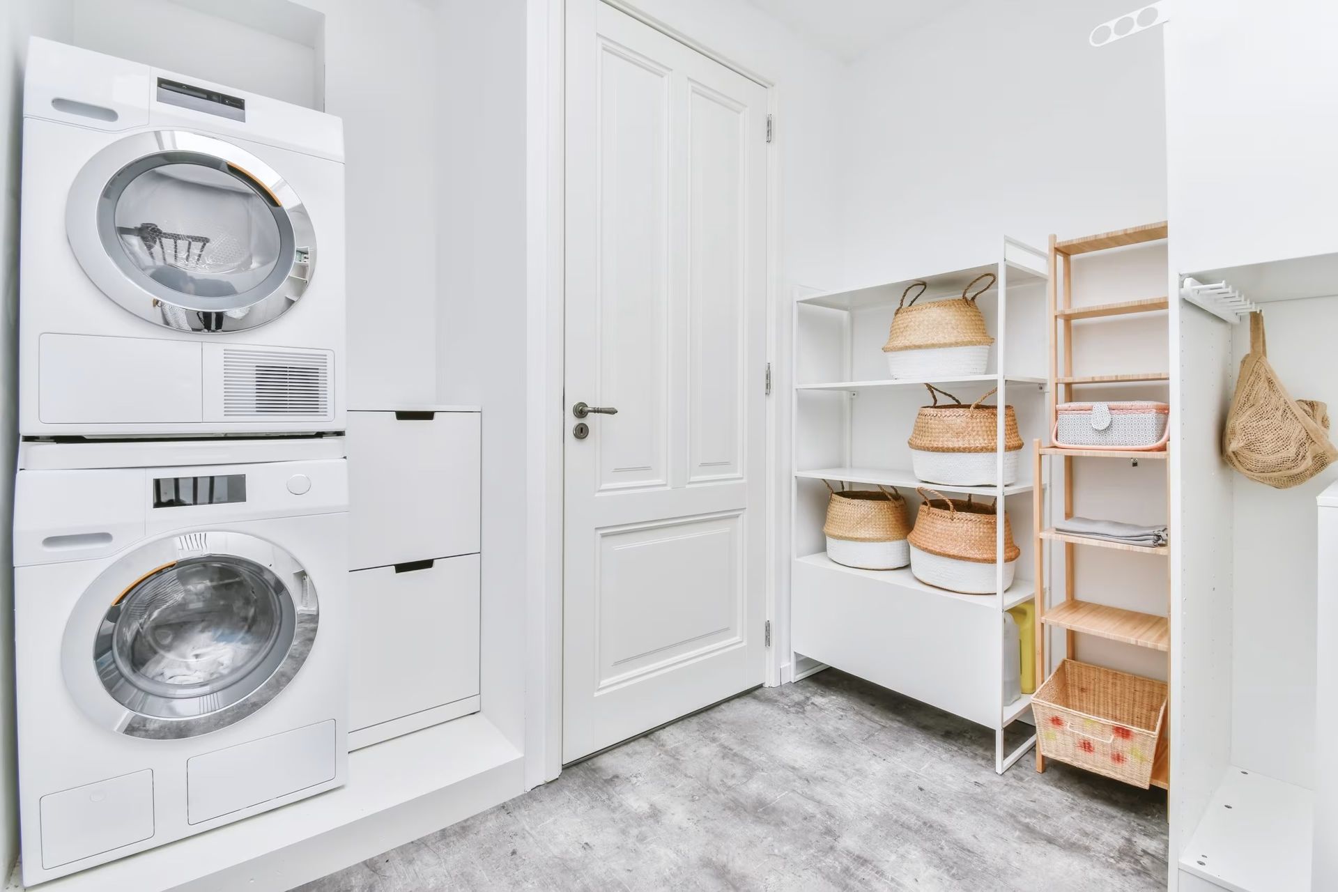 Bright laundry room with stacked washer and dryer, white door, shelves, and wicker baskets on gray floor.