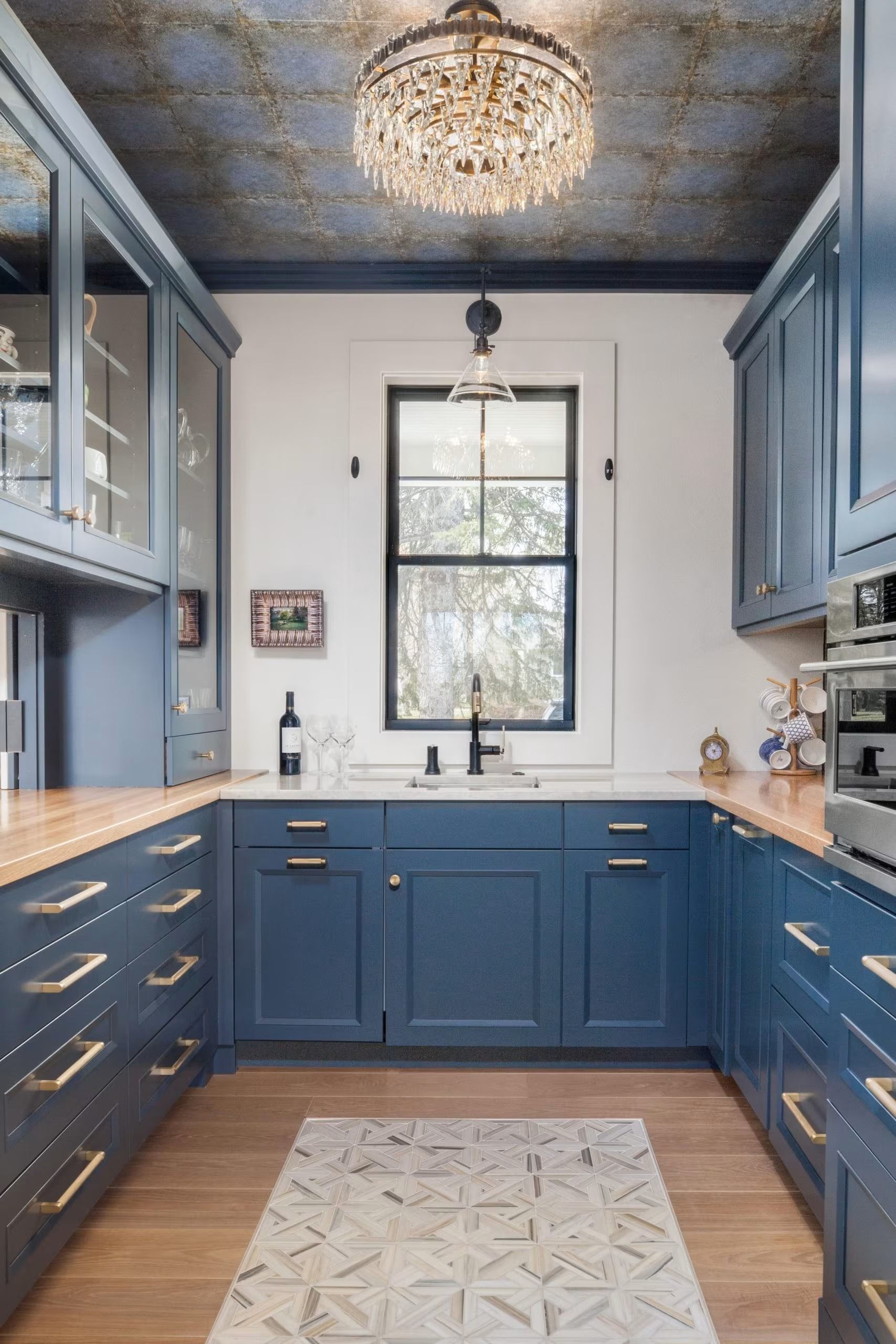 Blue galley kitchen with brass hardware, marble backsplash, and crystal chandelier