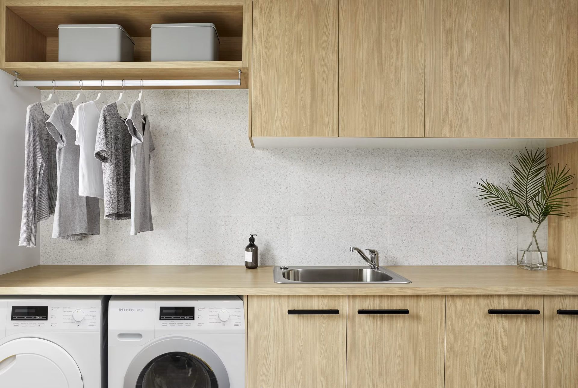 Modern laundry room with wood cabinets, washer and dryer, hanging clothes, and a countertop sink