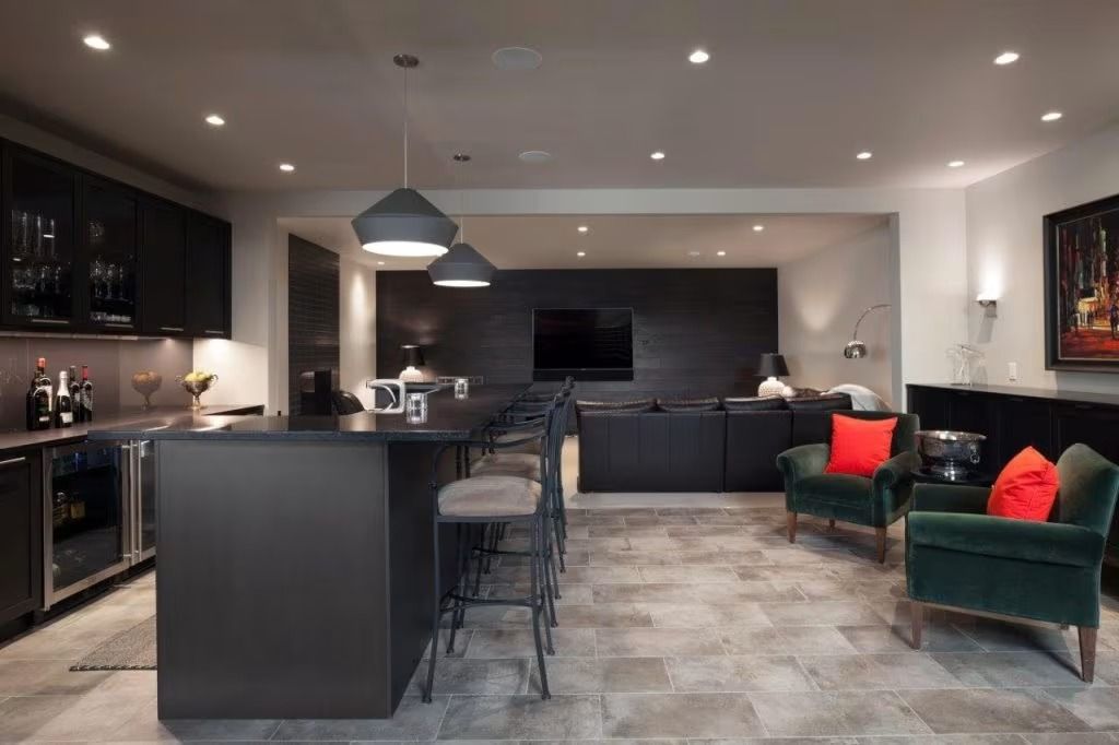 Modern open-plan kitchen and lounge with dark cabinets, gray floors, bar stools, and green chairs with red pillows