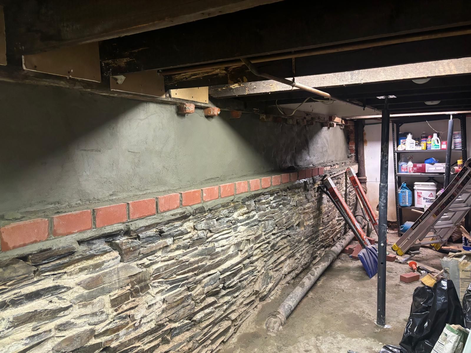Basement wall under construction: stone, brick, and gray cement, with beams and tools.