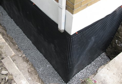 Black drainage membrane installed on a foundation wall, with gravel and a building corner.