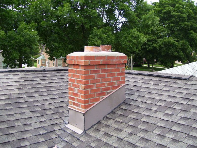 Brick chimney on a gray asphalt shingled roof with trees in the background.