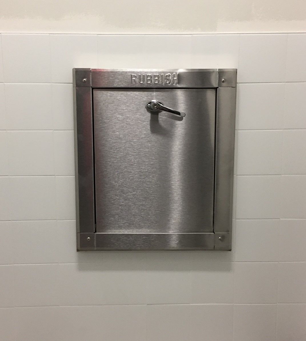 Stainless steel wall-mounted disposal door with handle, set in tiled wall. 