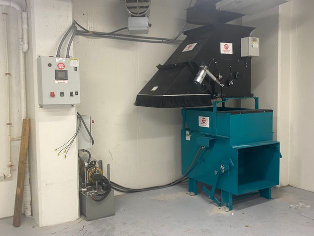 Industrial waste compactor and control panel in a corner, with blue and black machinery, gray conduit, and white walls.