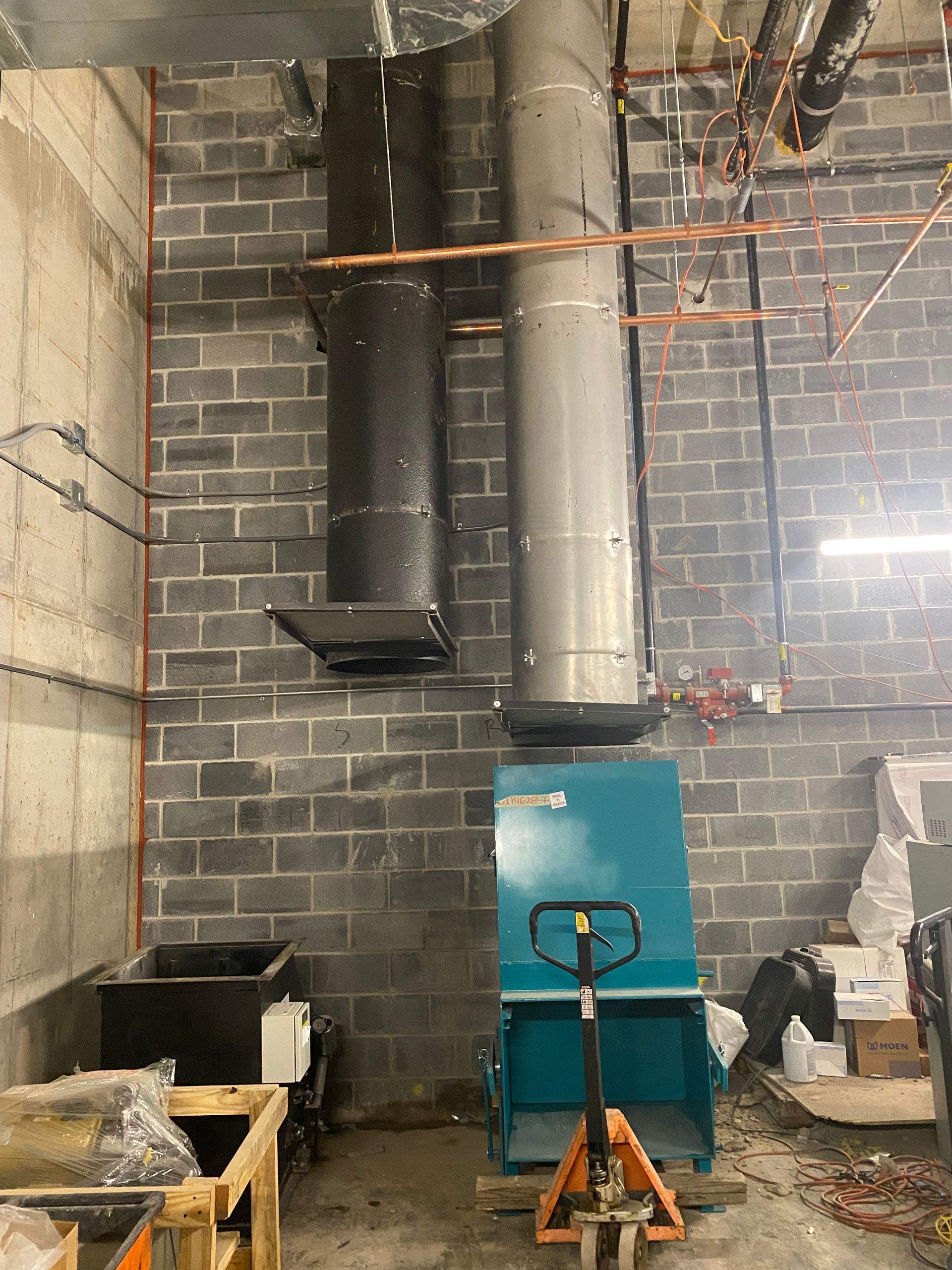Blue industrial machine with attached black and silver vent pipes against a gray brick wall.
