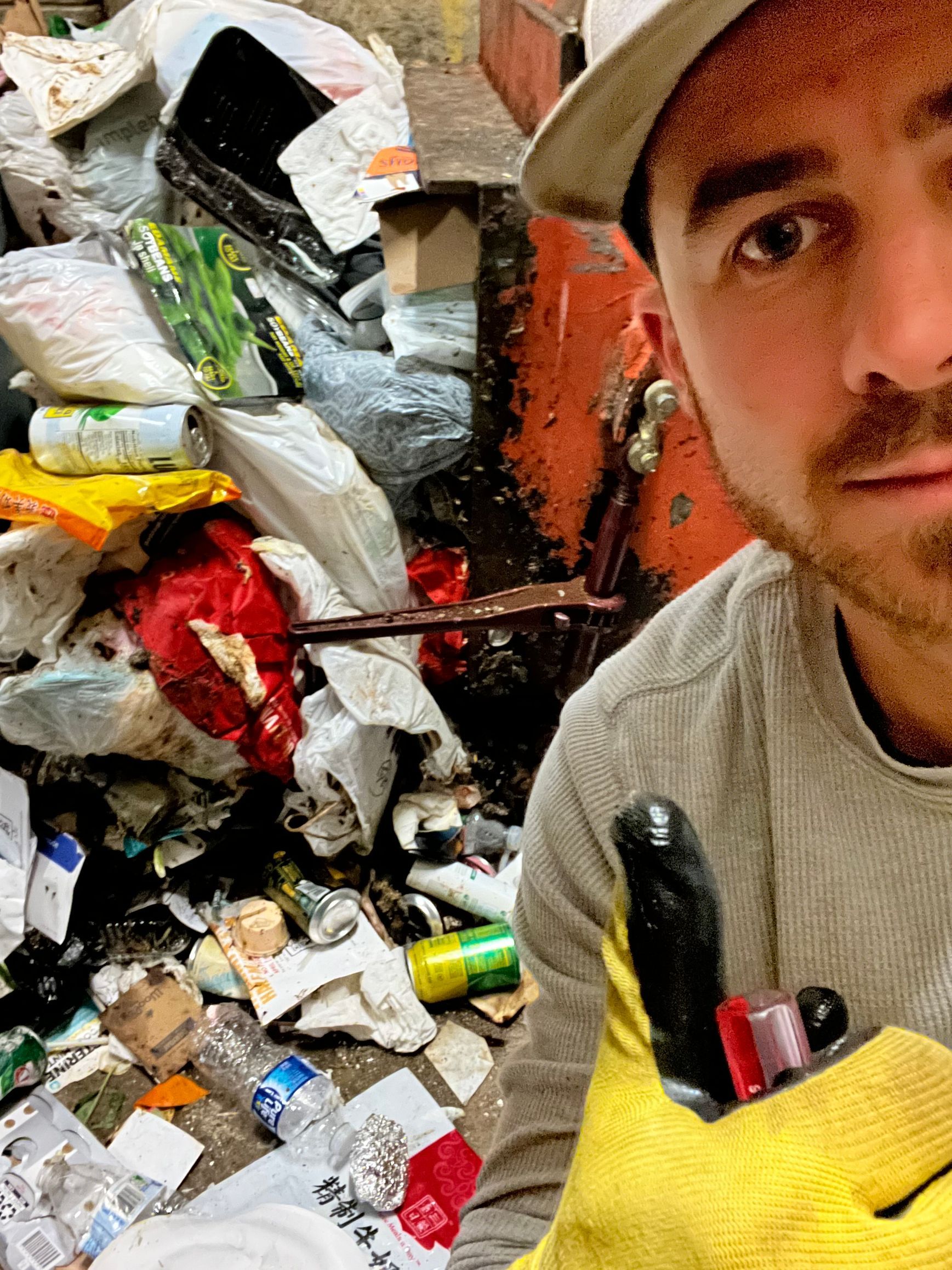 Man in cap and gloves takes selfie near a trash pile with colorful items.