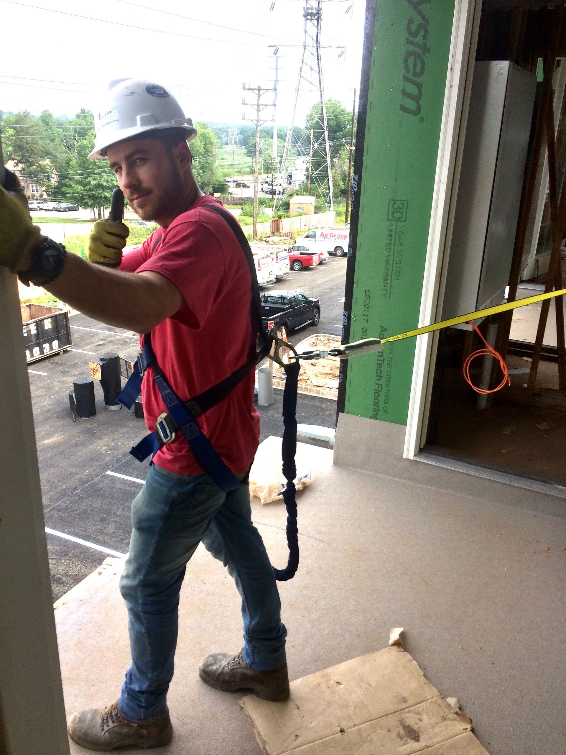 Construction worker in a safety harness giving a thumbs up. Outdoors, red shirt, hard hat, leaning against wall.