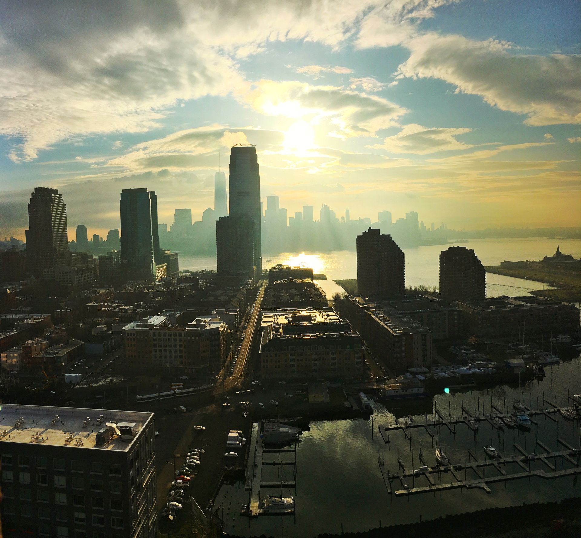Cityscape with sun breaking through clouds over water, high-rise buildings, docks and boats.