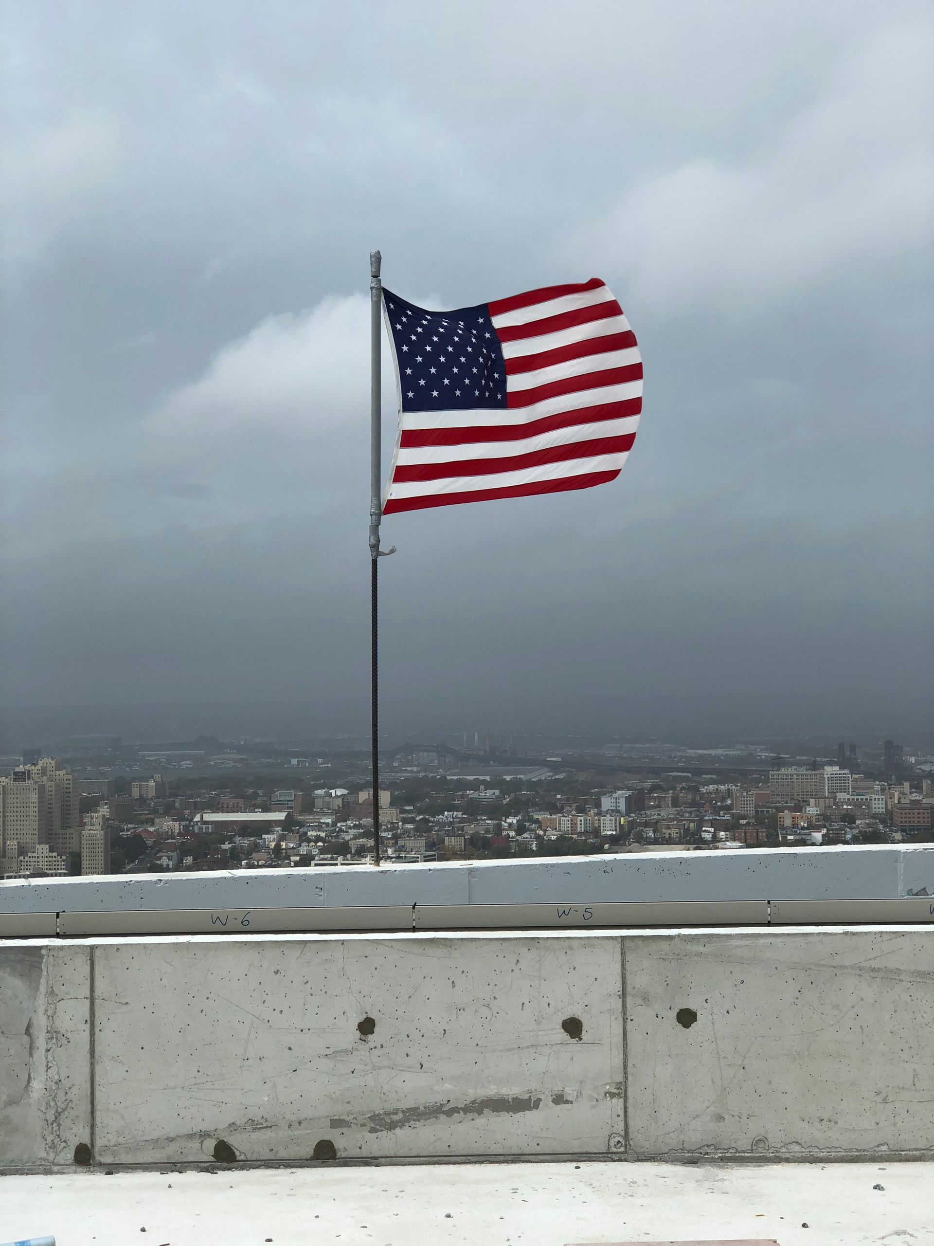 American flag waving on a pole against a cloudy sky, city in the distance.