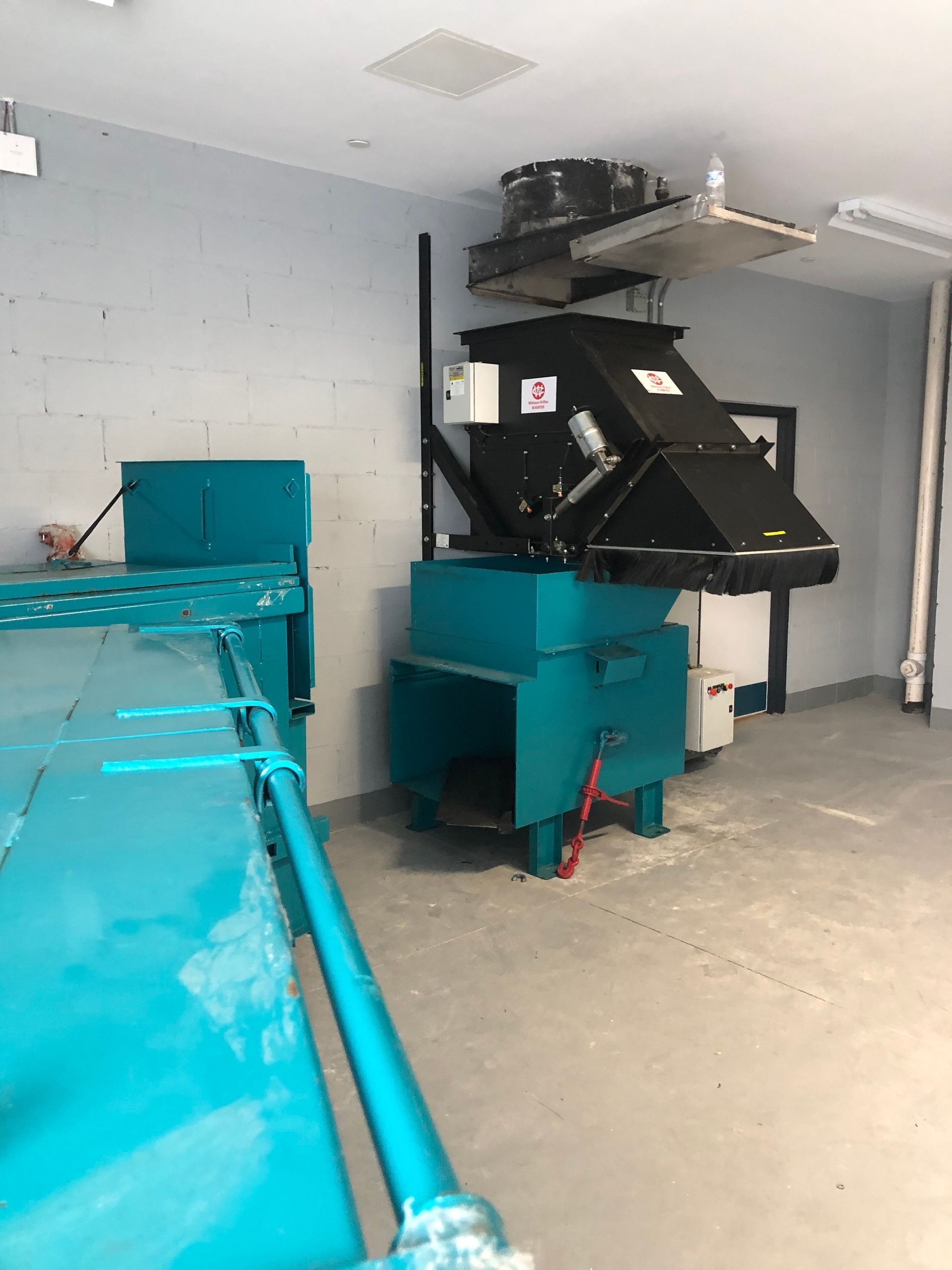 A teal trash compactor in a room with a black container. A blue table is in the left corner.