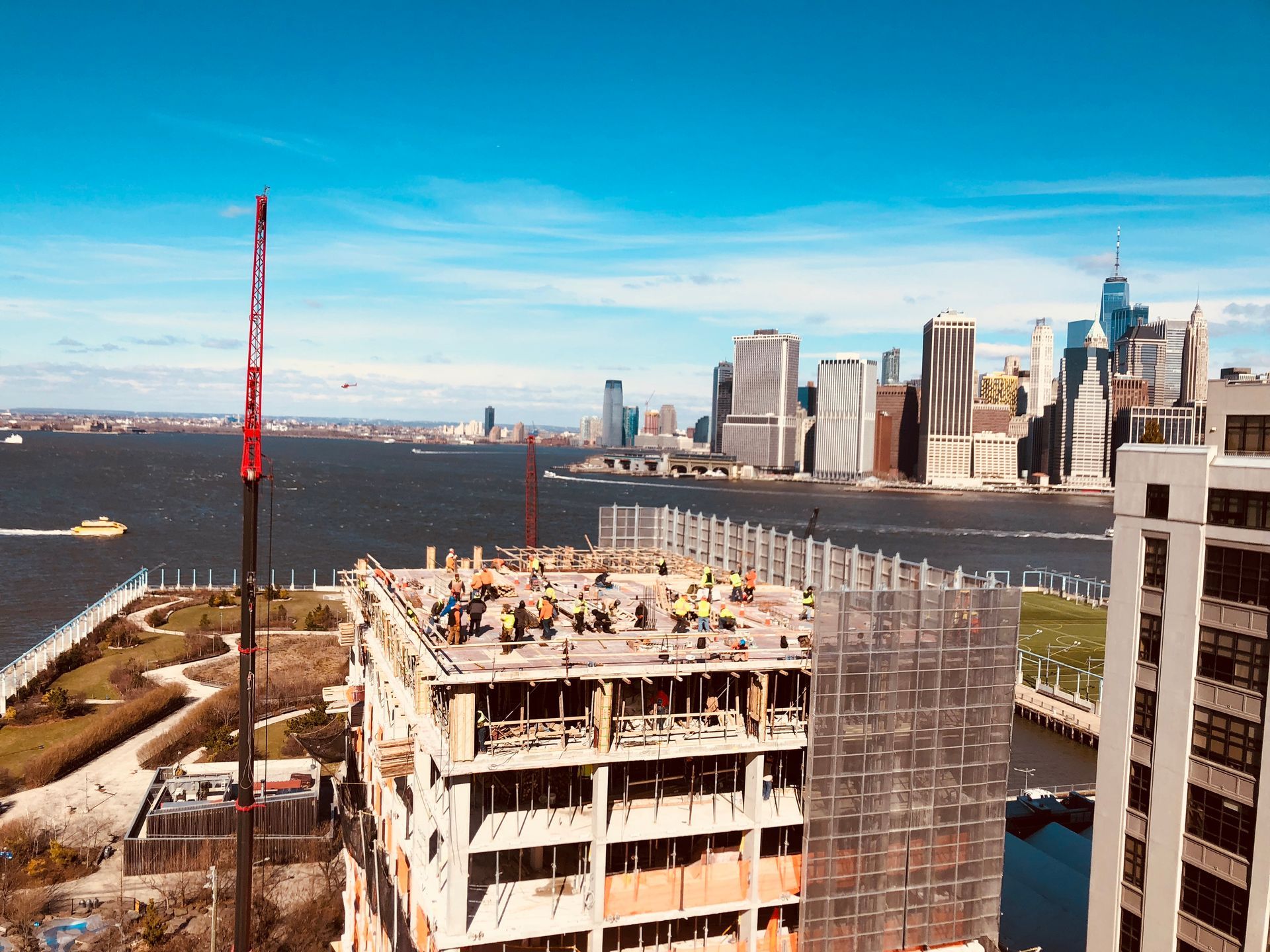 Construction site overlooking the water with workers, cranes, and NYC skyline on a bright, sunny day.