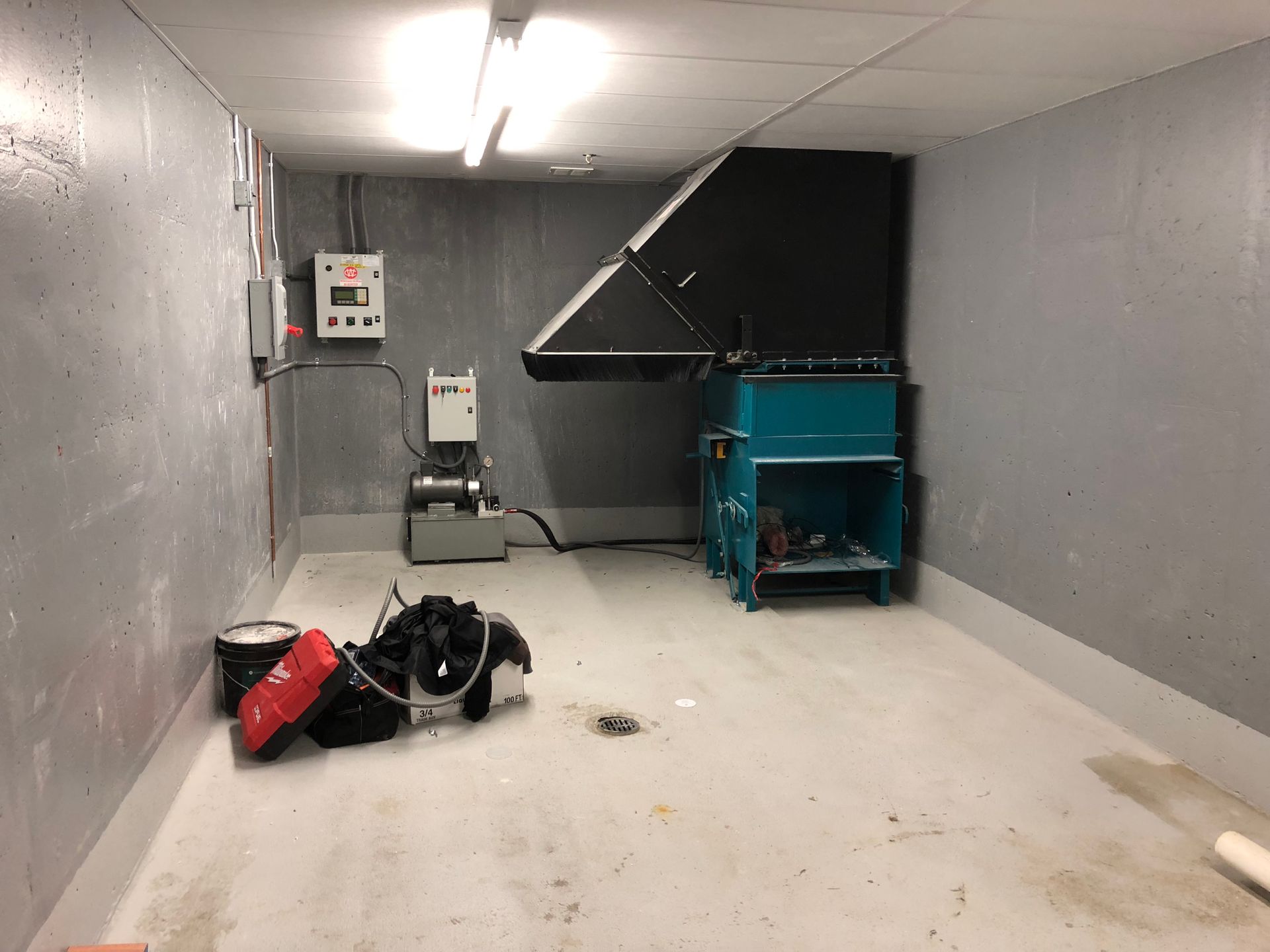A small industrial room with machinery, including a shredder and control panel. Walls are gray, and the floor is light.