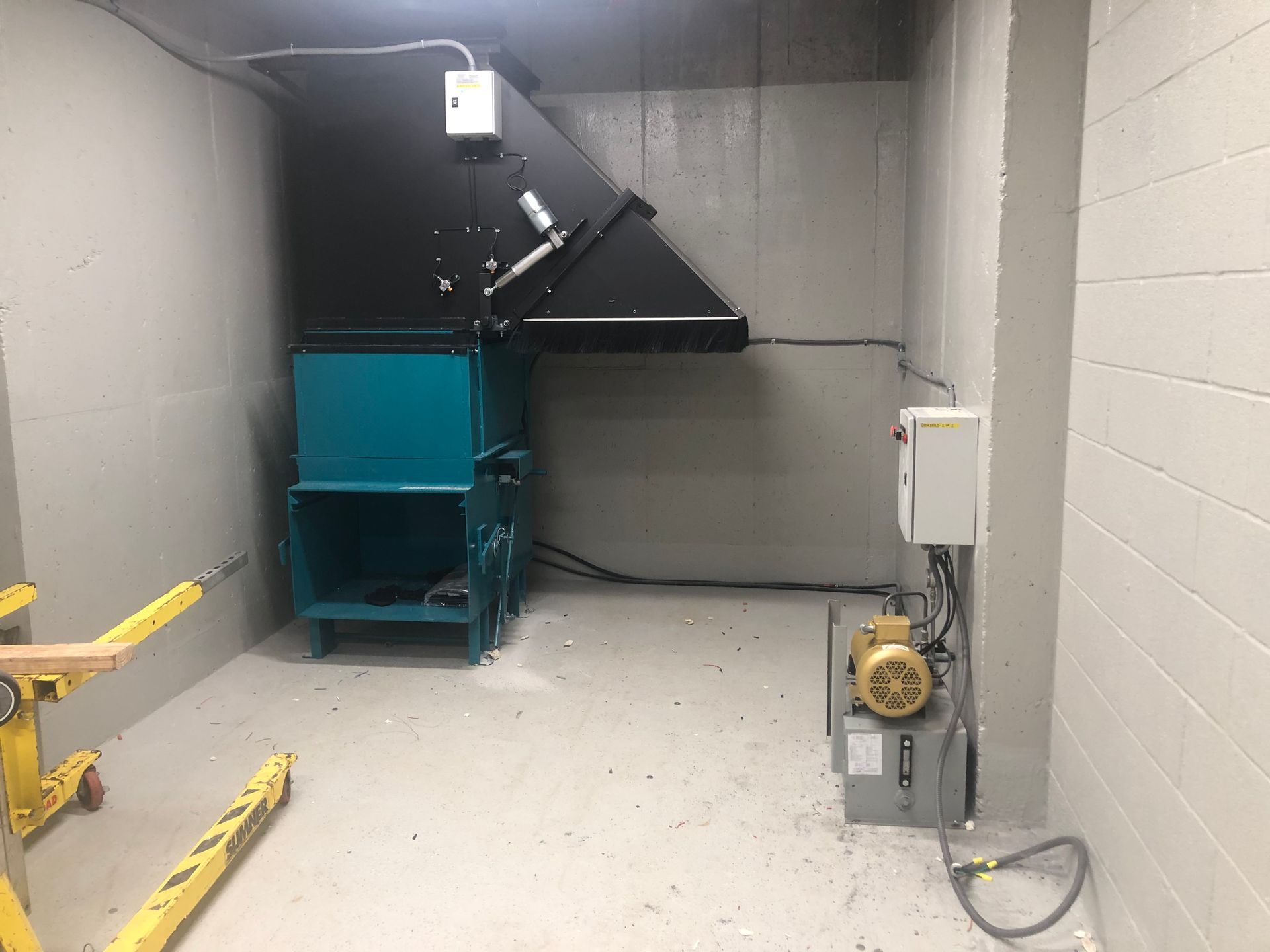 A large teal and black trash compactor in a concrete room with a forklift visible.