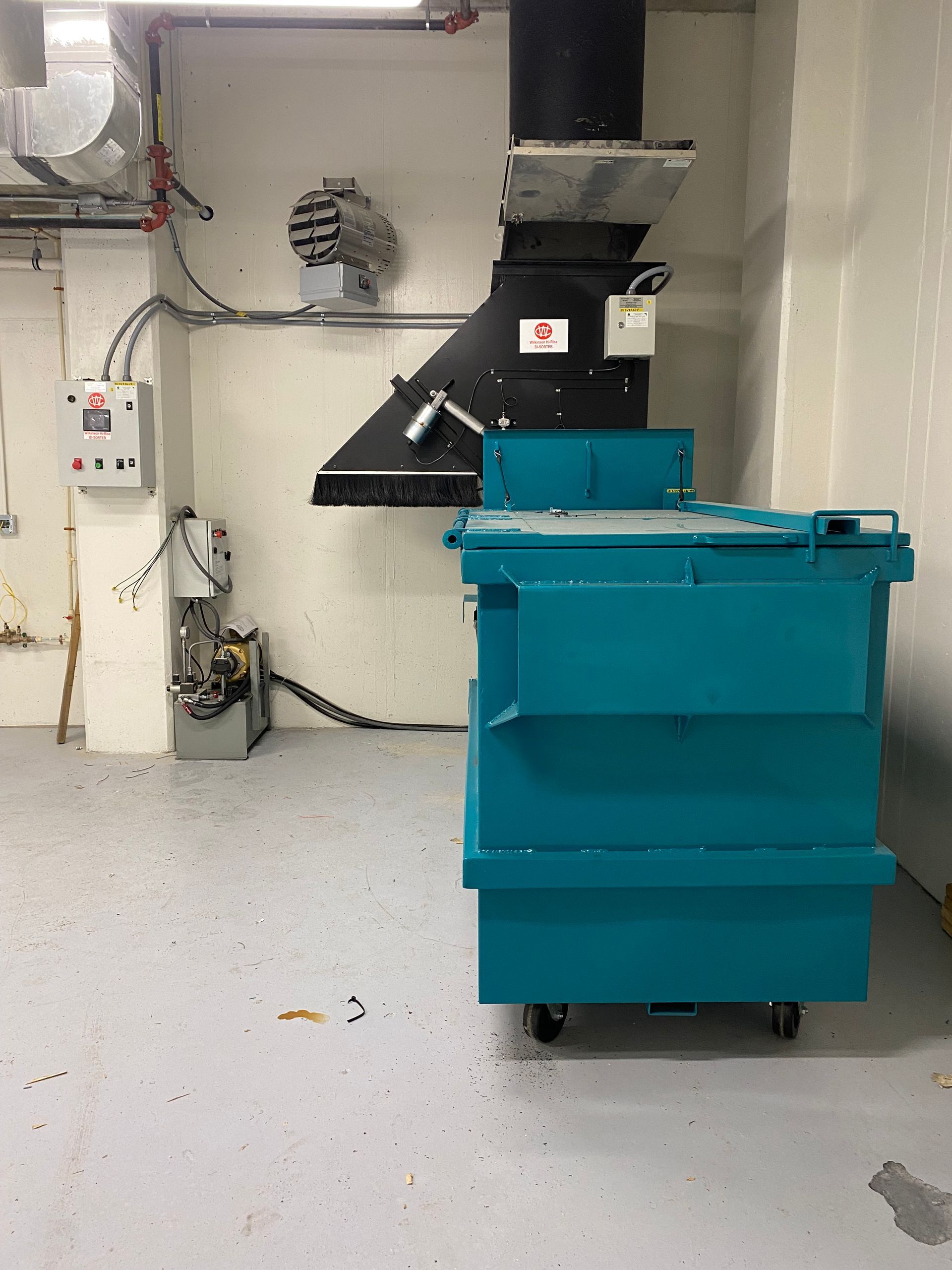 Blue trash compactor in a utility room. Black chute above the compactor.