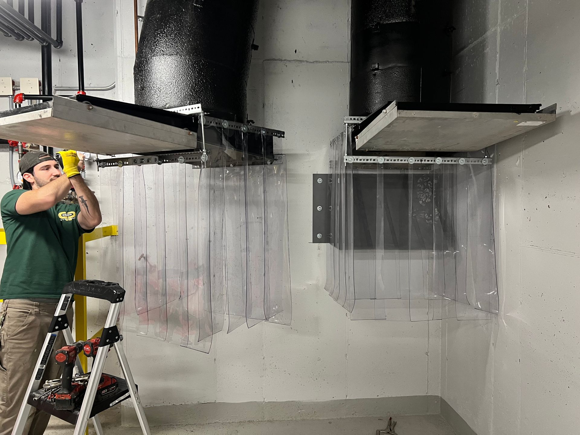 Man installs plastic curtain in industrial setting. He wears a green shirt and yellow gloves.  Gray and black infrastructure.