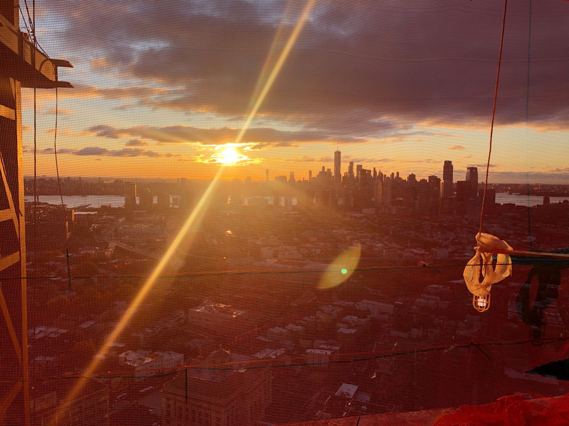 Sunset over city skyline, viewed through netting. Orange sun, dark buildings, golden light.