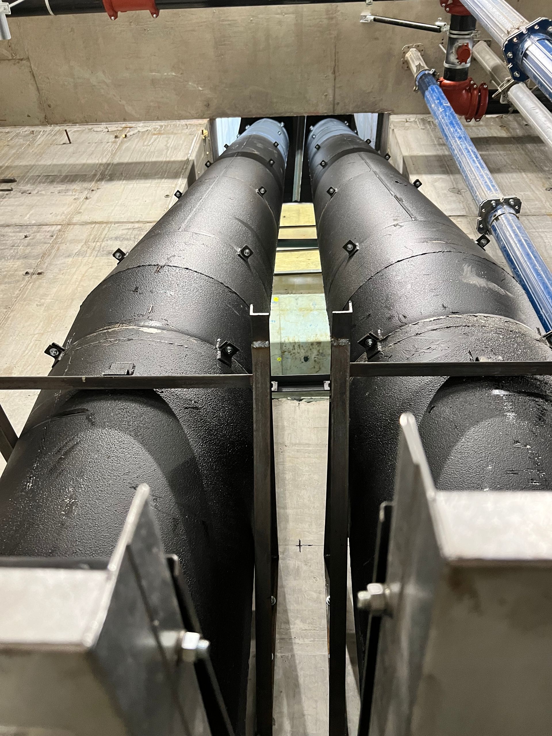 Two large insulated pipes running vertically between supports in a concrete space.