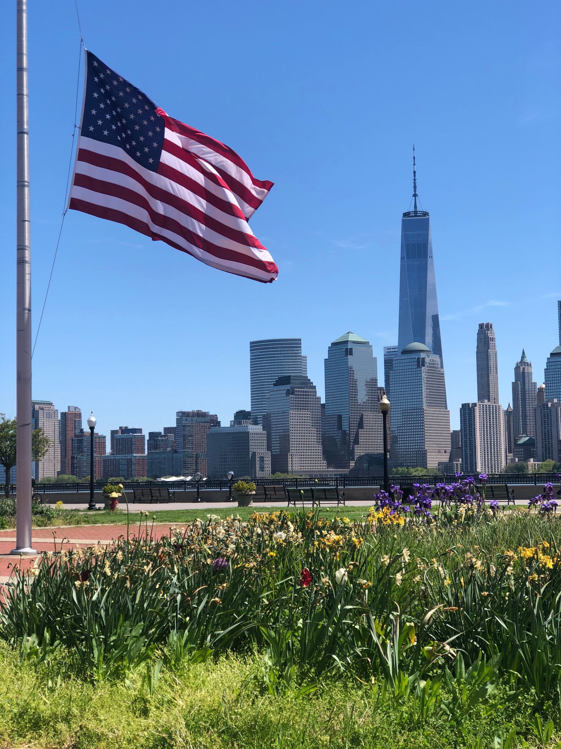American flag waving in front of the New York City skyline on a sunny day with flowers in the foreground.