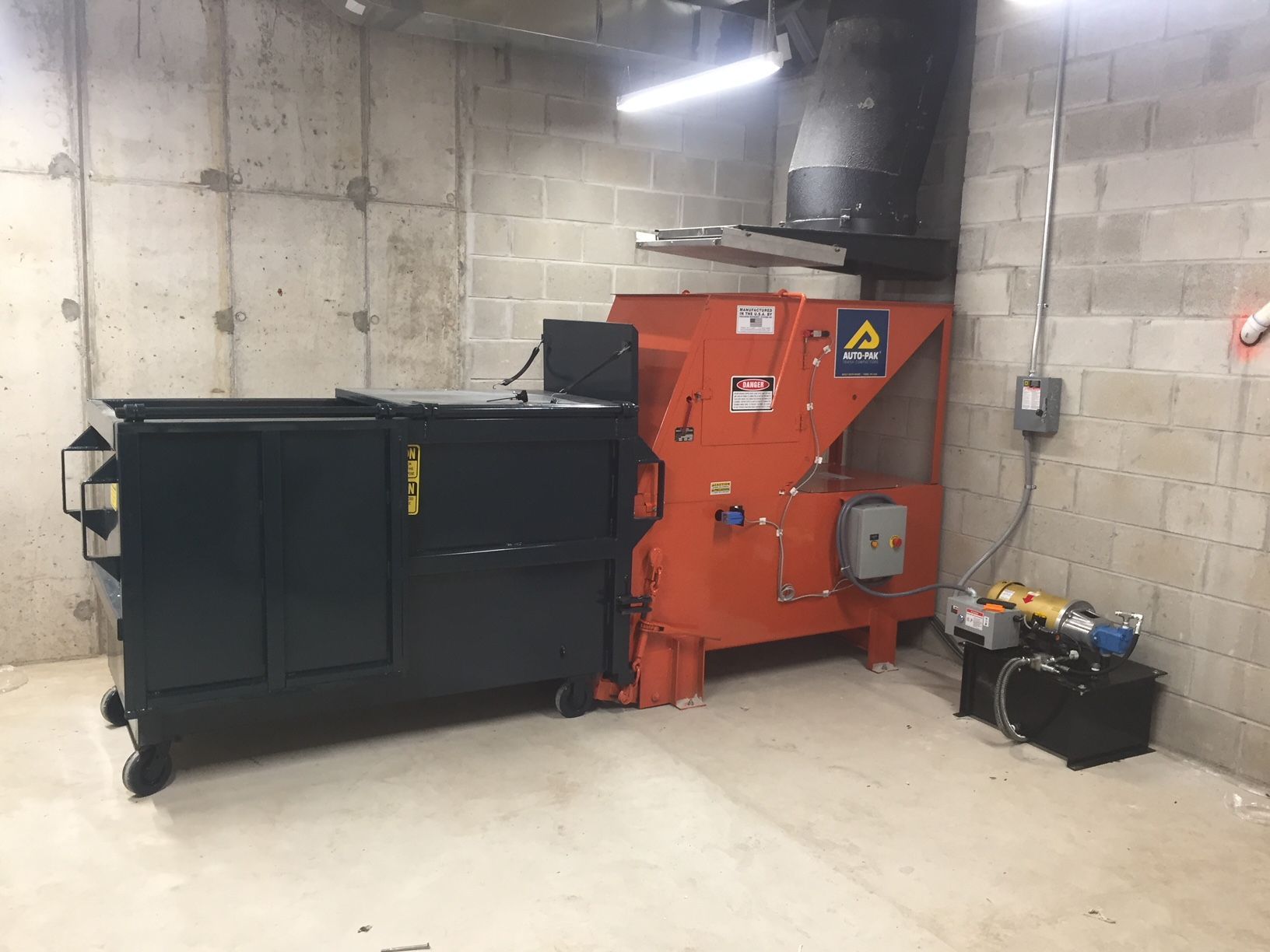 Orange and black trash compactor in a concrete room with a hydraulic pump.