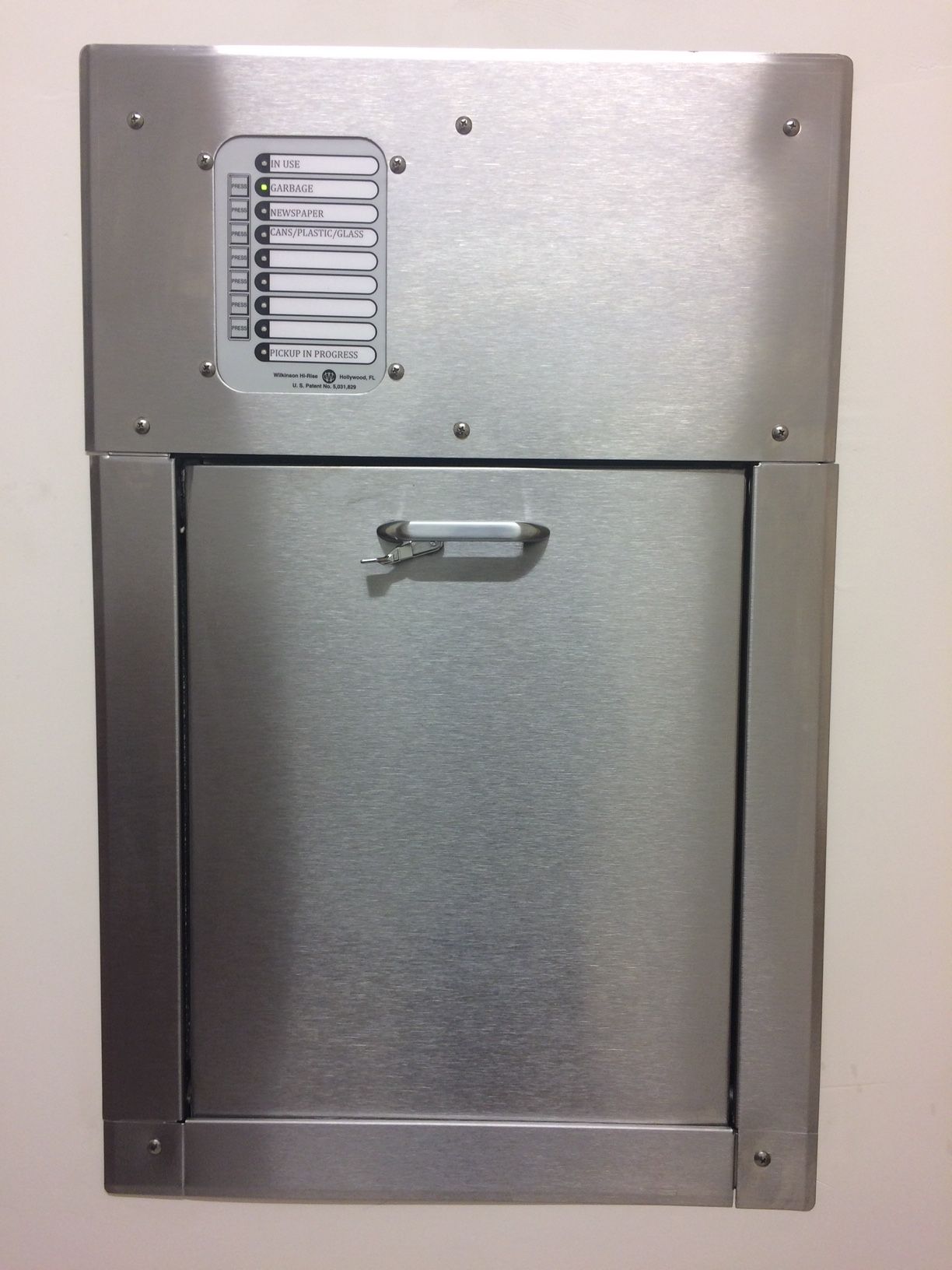 Stainless steel wall-mounted trash receptacle with a hinged door and a latch. Includes a label with instructions.